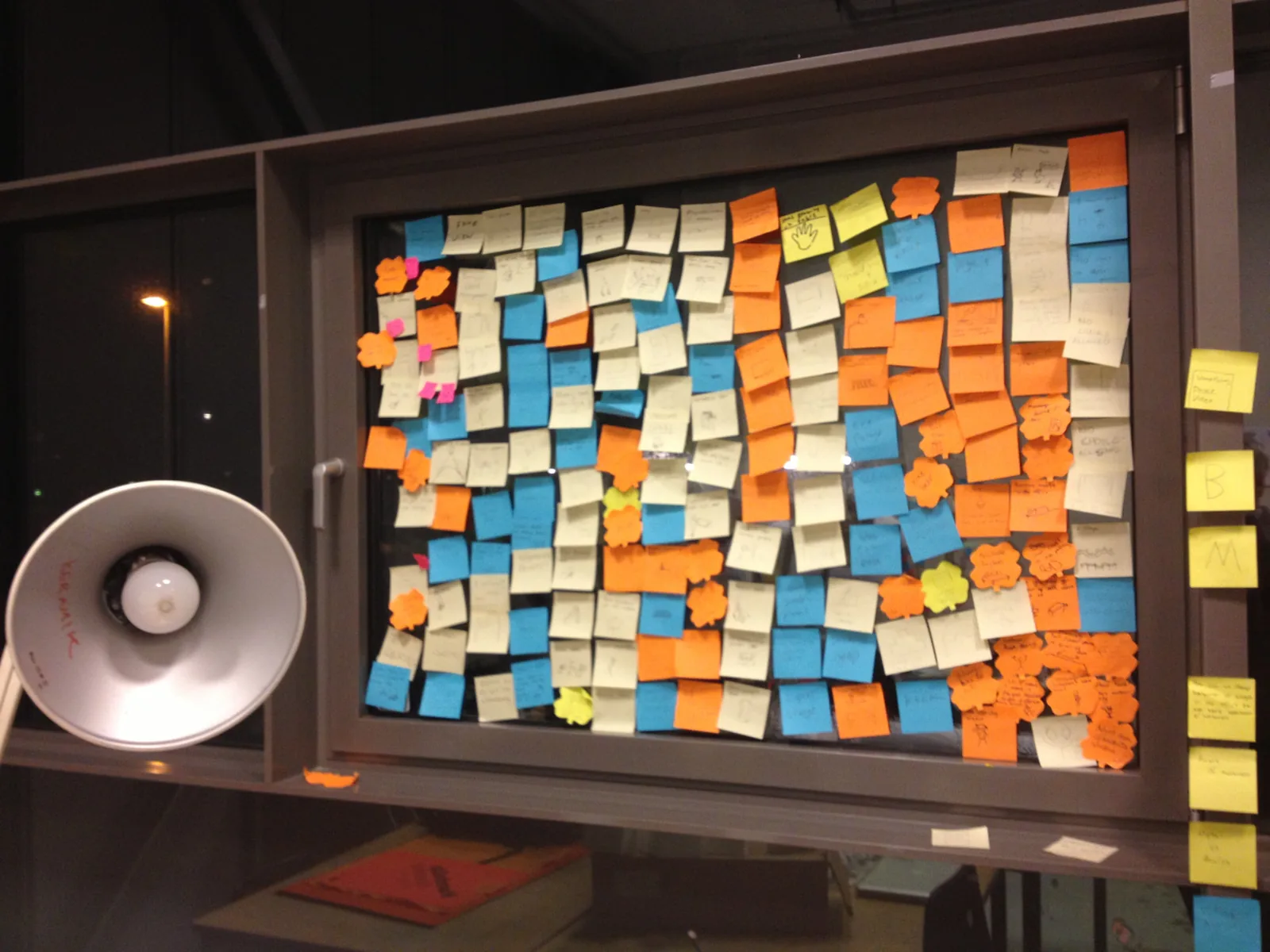 Glass wall covered with handwritten sticky notes from a design workshop, lit by an angled work lamp